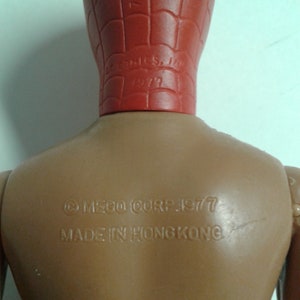 Op de afbeelding: Een bruin plastic actiefiguur van Spider-Man met een rood hoofd en een webdesign. Op de achterkant van het figuur staat de tekst "&copy; MEGO CORP. 1977 MADE IN HONG KONG".
