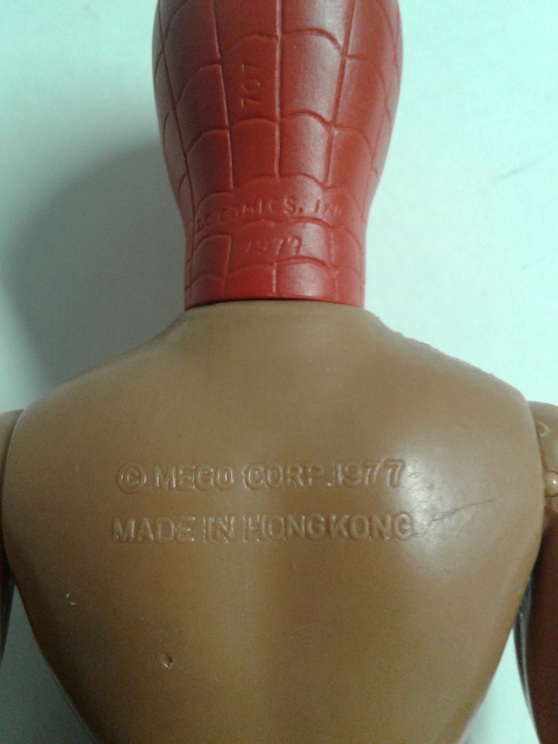 Op de afbeelding: Een bruin plastic actiefiguur van Spider-Man met een rood hoofd en een webdesign. Op de achterkant van het figuur staat de tekst "&copy; MEGO CORP. 1977 MADE IN HONG KONG".