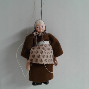Könnte beinhalten: Eine braune Filzpuppe einer älteren Frau mit weißem Gesicht, blauen Augen und einer braunen Schürze mit weißem Muster. Die Puppe trägt ein braunes Filzkleid und ein weißes Kopftuch mit braunem und weißem Muster.