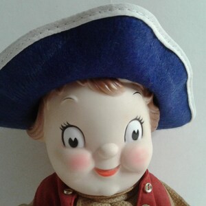 Pu&ograve; includere: Una bambola vintage con un viso sorridente, guance rosee e grandi occhi. La bambola indossa un cappello blu con finiture bianche e un gilet rosso sopra un maglione color cachi. I lineamenti della bambola sono dipinti con dettagli.