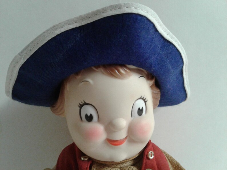 Pu&ograve; includere: Una bambola vintage con un viso sorridente, guance rosee e grandi occhi. La bambola indossa un cappello blu con finiture bianche e un gilet rosso sopra un maglione color cachi. I lineamenti della bambola sono dipinti con dettagli.