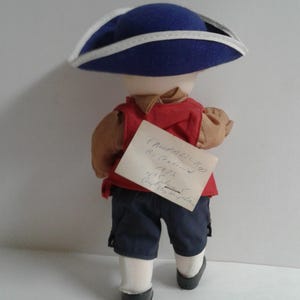 Pu&ograve; includere: Bambola vestita con abito storico, vista da dietro. Indossa un cappello blu con finiture bianche, un gilet rosso, pantaloncini blu navy e scarpe nere. Una nota scritta a mano &egrave; attaccata alla schiena della bambola.