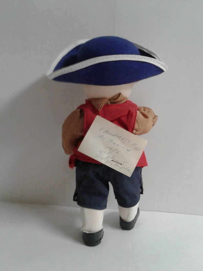 Pu&ograve; includere: Bambola vestita con abito storico, vista da dietro. Indossa un cappello blu con finiture bianche, un gilet rosso, pantaloncini blu navy e scarpe nere. Una nota scritta a mano &egrave; attaccata alla schiena della bambola.