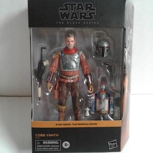 May include: A Star Wars: The Mandalorian action figure of Cobb Vanth, a bounty hunter from the planet Tatooine. The figure is wearing a brown leather jacket, a red and brown vest, and a silver helmet. The figure is holding a blaster rifle in his right hand and a blaster pistol in his left hand. The figure is standing on a black base with the Star Wars: The Black Series logo.
