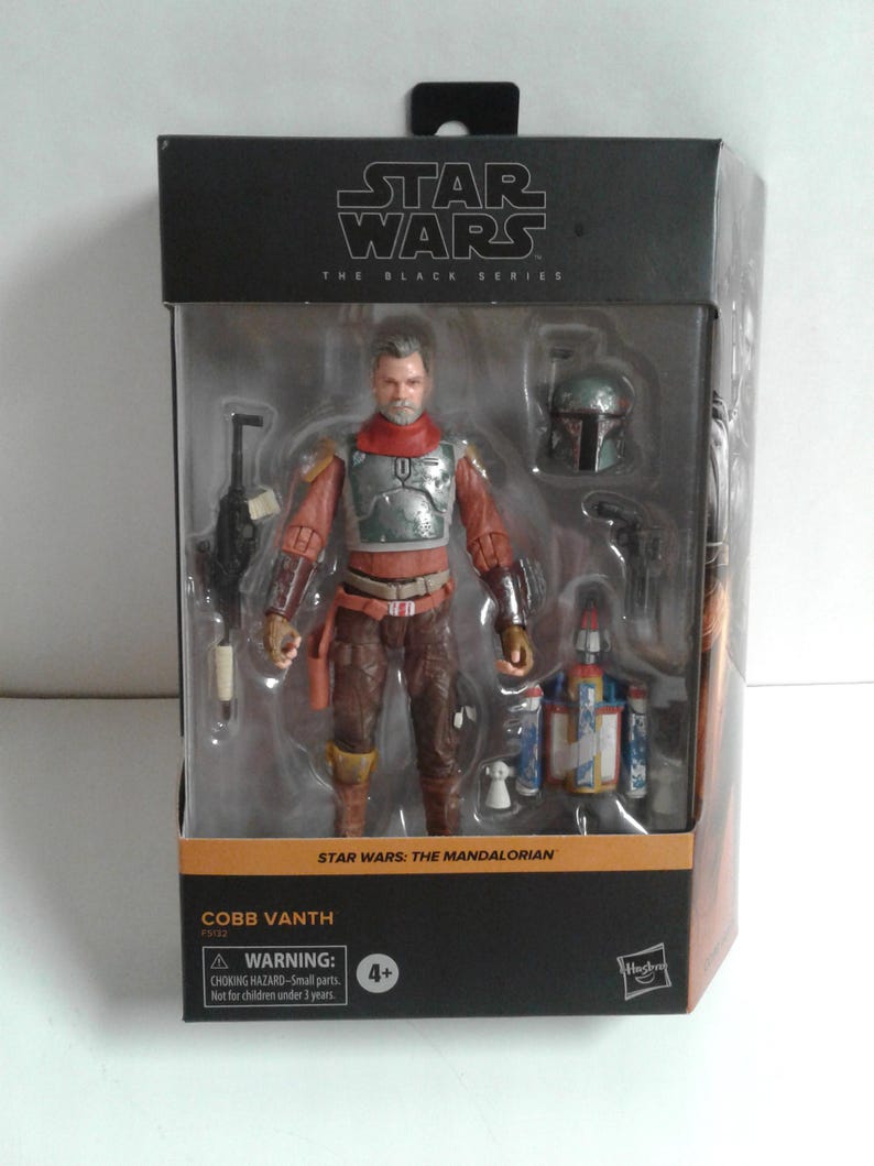 May include: A Star Wars: The Mandalorian action figure of Cobb Vanth, a bounty hunter from the planet Tatooine. The figure is wearing a brown leather jacket, a red and brown vest, and a silver helmet. The figure is holding a blaster rifle in his right hand and a blaster pistol in his left hand. The figure is standing on a black base with the Star Wars: The Black Series logo.