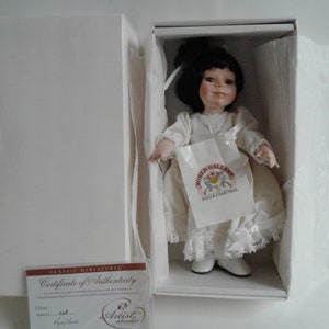 Pode incluir: Uma boneca de porcelana com cabelo preto e um vestido branco. A boneca est&aacute; usando sapatos brancos e segurando uma bolsa branca com o texto "World Gallery Dolls & Collectibles". A boneca est&aacute; em uma caixa branca com um certificado de autenticidade que diz "Classic Miniatures Certificate of Authenticity China Edition # 548 Artist Studio Lee Middleton Original Dolls, Inc."