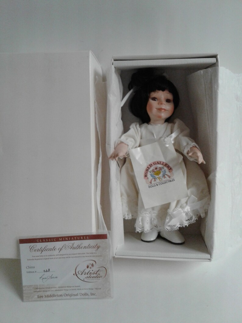 Pode incluir: Uma boneca de porcelana com cabelo preto e um vestido branco. A boneca est&aacute; usando sapatos brancos e segurando uma bolsa branca com o texto "World Gallery Dolls & Collectibles". A boneca est&aacute; em uma caixa branca com um certificado de autenticidade que diz "Classic Miniatures Certificate of Authenticity China Edition # 548 Artist Studio Lee Middleton Original Dolls, Inc."