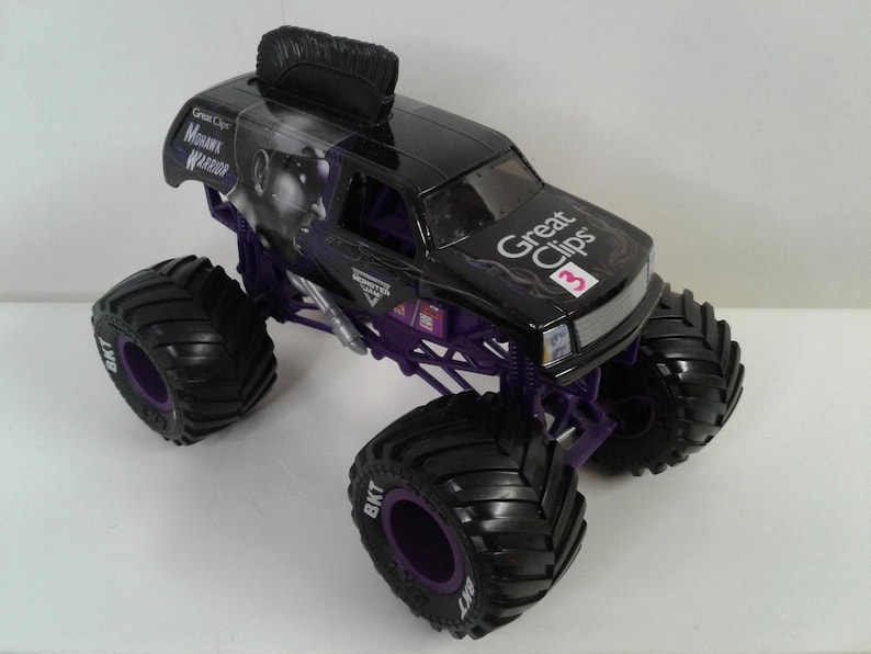 May include: Black and purple toy monster truck with large tyres. The truck features the text "Great Clips" and "Mohawk Warrior". The tyres have the text "BKT". The truck has a black mohawk on top.