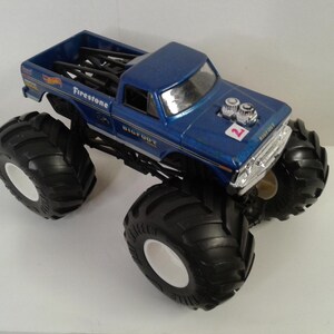 May include: A blue toy monster truck with large black tyres and white rims. The truck has the word "Firestone" and "BIGFOOT" printed on it. The number "2" is visible on the bonnet. The truck bed contains black bars.
