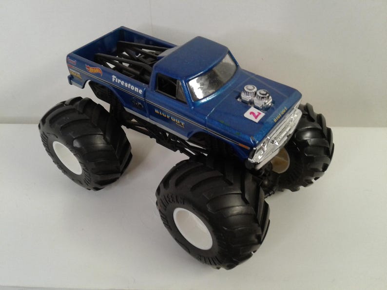 May include: A blue toy monster truck with large black tyres and white rims. The truck has the word "Firestone" and "BIGFOOT" printed on it. The number "2" is visible on the bonnet. The truck bed contains black bars.