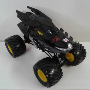 May include: Black monster truck toy with large black tyres and yellow rims. The truck has a Batman design with the Batman logo and the words "HA HA" in red. The truck has a number 6 on the side.