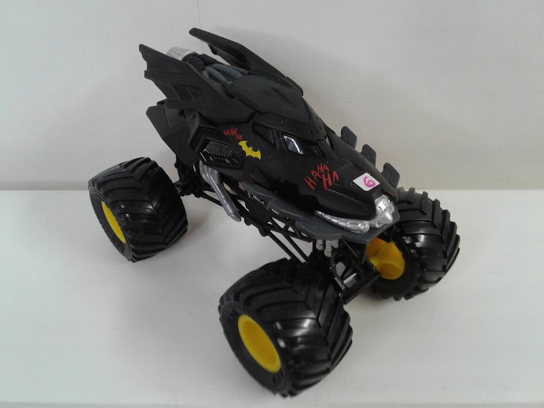 May include: Black monster truck toy with large black tyres and yellow rims. The truck has a Batman design with the Batman logo and the words "HA HA" in red. The truck has a number 6 on the side.