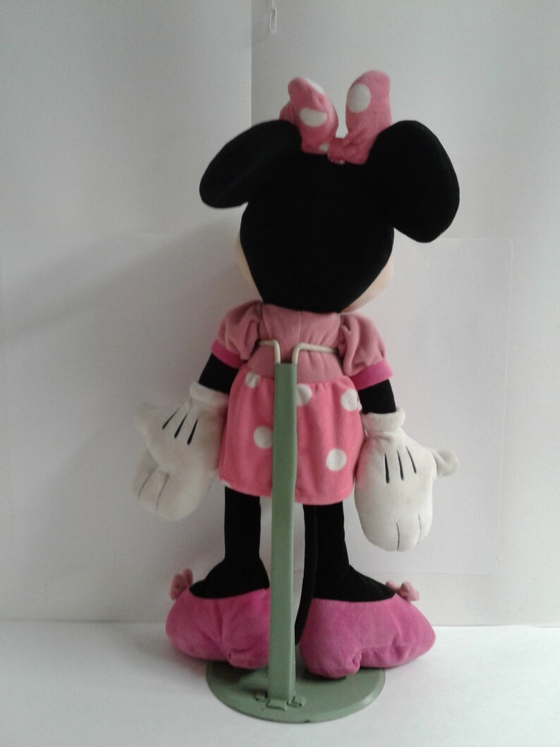 May include: A pink and white polka dot dress with a black bow on top of the head. The plush toy is standing on a green stand.