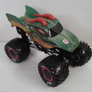 May include: A toy monster truck with a dragon design in green and red. The truck has large black tyres with red detailing and the number 5 on the bonnet. The dragon's head is green with red horns and yellow eyes.