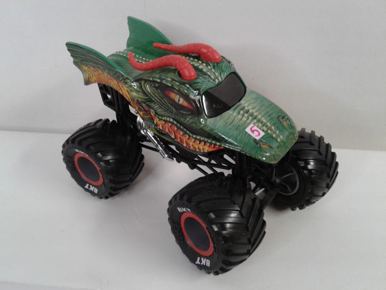 May include: A toy monster truck with a dragon design in green and red. The truck has large black tyres with red detailing and the number 5 on the bonnet. The dragon's head is green with red horns and yellow eyes.