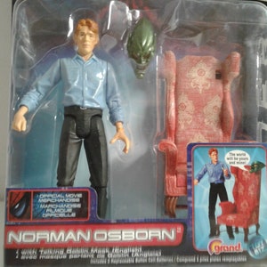 May include: Norman Osborn action figure from the Spider-Man movie. The figure is wearing a blue shirt, black pants, and a red tie. The figure is holding a green mask in his right hand. The figure is standing in front of a red chair. The packaging includes the text "The world will be yours and mine!"