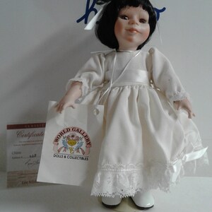 Pode incluir: Uma boneca de porcelana com cabelo preto e um vestido branco. A boneca est&aacute; usando sapatos brancos e tem um la&ccedil;o branco no cabelo. A boneca est&aacute; segurando uma etiqueta branca que diz "World Gallery Dolls & Collectibles".