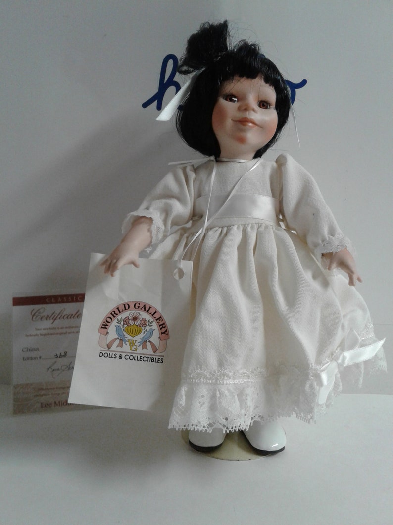 Pode incluir: Uma boneca de porcelana com cabelo preto e um vestido branco. A boneca est&aacute; usando sapatos brancos e tem um la&ccedil;o branco no cabelo. A boneca est&aacute; segurando uma etiqueta branca que diz "World Gallery Dolls & Collectibles".
