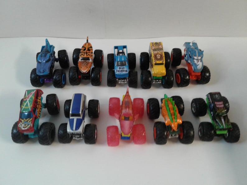 Może przedstawiać: Kolekcja zabawkowych monster truck&oacute;w w r&oacute;żnych kolorach, w tym niebieskim, pomarańczowym, ż&oacute;łtym i zielonym. Każdy truck ma duże, czarne opony i unikalne wzory, takie jak paski tygrysa i płetwy rekina. Ciężar&oacute;wki są ułożone na białej powierzchni.