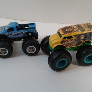 Może przedstawiać: Dwa zabawki monster truck. Jeden jest jasnoniebieski z napisem "BLUE THUNDER" z boku. Drugi jest ż&oacute;łty z napisem "HOUND". Oba mają duże czarne opony.