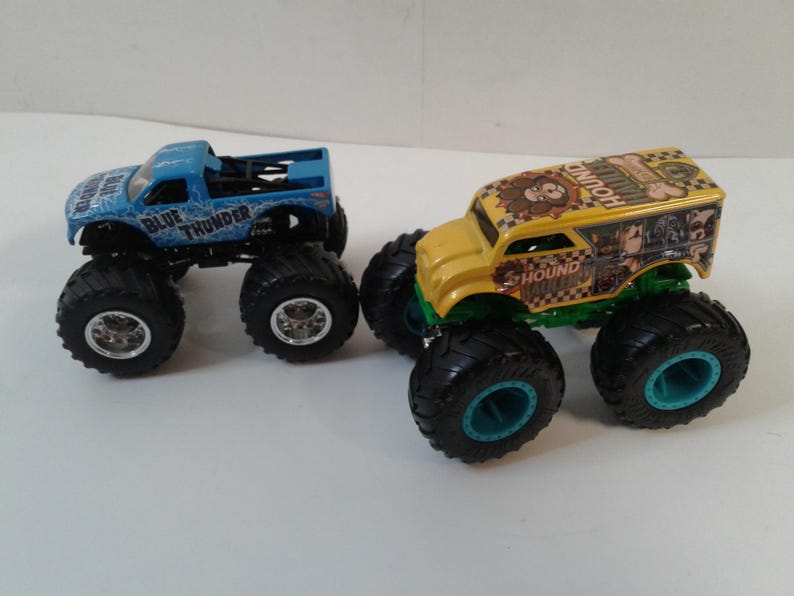 Może przedstawiać: Dwa zabawki monster truck. Jeden jest jasnoniebieski z napisem "BLUE THUNDER" z boku. Drugi jest ż&oacute;łty z napisem "HOUND". Oba mają duże czarne opony.