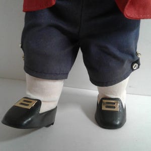 Pu&ograve; includere: Gambe di bambola vestite con pantaloncini blu navy con bottoni decorativi, calzini bianchi e scarpe nere con fibbie color oro. La giacca rossa della bambola &egrave; visibile nella parte superiore del fotogramma.