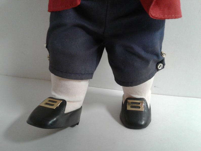 Pu&ograve; includere: Gambe di bambola vestite con pantaloncini blu navy con bottoni decorativi, calzini bianchi e scarpe nere con fibbie color oro. La giacca rossa della bambola &egrave; visibile nella parte superiore del fotogramma.