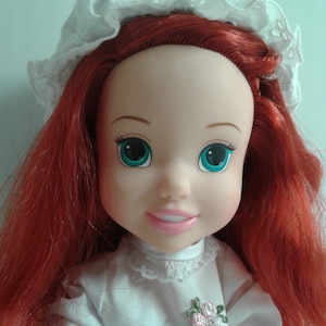 K&ouml;nnte beinhalten: Eine rothaarige Puppe mit einem wei&szlig;en Spitzenh&auml;ubchen und einem wei&szlig;en Kleid mit Spitzenbesatz und einer floralen Verzierung. Die Puppe hat blaue Augen und ein rosafarbenes L&auml;cheln.