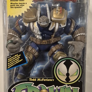 Op de afbeelding: Een Badrock actiefiguur van Todd Toys in de originele verpakking. Het figuur is blauw en bruin met schouder-silo's die raketten afvuren. De verpakking bevat het Spawn-logo en de tekst Missiles Fire from Shoulder Silos en Ages 4 and Up.