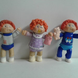 Può includere: Tre bambole Cabbage Patch Kids con i capelli rossi, vestite con abiti diversi. La bambola a sinistra tiene un cono gelato giocattolo, quella al centro indossa un vestito viola e tiene una coperta rosa, e quella a destra indossa un maglione blu con un motivo a rombi e tiene un orsacchiotto rosa.