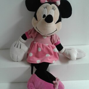 May include: A large plush Minnie Mouse toy wearing a pink dress with white polka dots and pink shoes. She has a black nose and large black eyes.