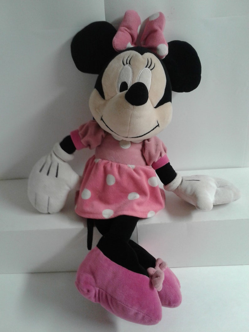 May include: A large plush Minnie Mouse toy wearing a pink dress with white polka dots and pink shoes. She has a black nose and large black eyes.