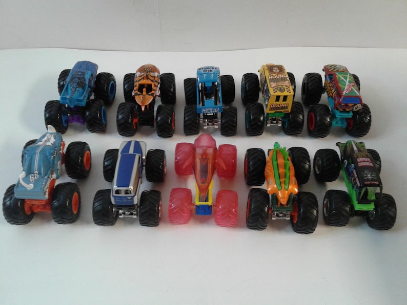 Może przedstawiać: Kolekcja zabawkowych monster truck&oacute;w w r&oacute;żnych kolorach, w tym niebieskim, pomarańczowym, zielonym i ż&oacute;łtym. Każdy truck ma duże, czarne opony i unikalne wzory na nadwoziu. Ciężar&oacute;wki są ułożone w rzędach na białej powierzchni.