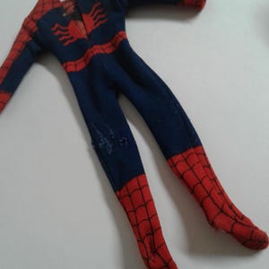 Op de afbeelding: Een blauw en rood Spiderman-kostuum voor een pop. Het kostuum heeft een spinnenwebontwerp en een spinnenlogo op de borst.