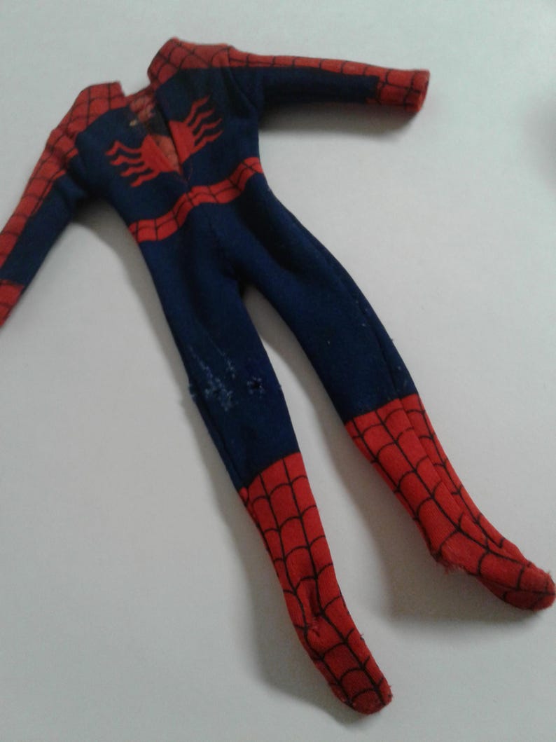 Op de afbeelding: Een blauw en rood Spiderman-kostuum voor een pop. Het kostuum heeft een spinnenwebontwerp en een spinnenlogo op de borst.