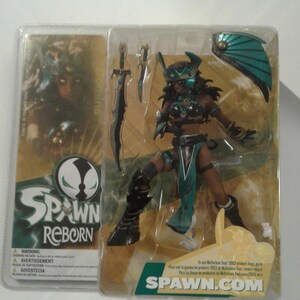 May include: A Spawn Reborn action figure in original blister packaging. The figure features a dark-skinned warrior wearing teal and black armour, a helmet, and a loincloth. Accessories include a long sword, a dagger, and a teal shield. The packaging displays the Spawn Reborn logo and the website address Spawn.com.