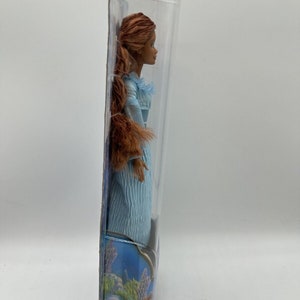 May include: A doll in a blue dress with brown hair in a ponytail. The doll is packaged in a clear plastic box with a blue and purple background.
