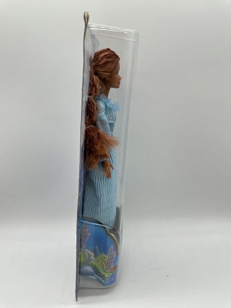 May include: A doll in a blue dress with brown hair in a ponytail. The doll is packaged in a clear plastic box with a blue and purple background.