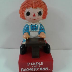 May include: A vintage Raggedy Ann stapler. The stapler has a red base with the text "STAPLE WITH RAGGEDY ANN". The stapler features a Raggedy Ann doll figure on top, with orange hair, a blue dress, and a white apron.
