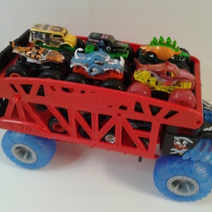 Może przedstawiać: Czerwona zabawka ciężar&oacute;wki potwora Hot Wheels przewożąca kilka mniejszych ciężar&oacute;wek potwor&oacute;w. Duża ciężar&oacute;wka ma niebieskie koła i czarno-czerwony korpus. Mniejsze ciężar&oacute;wki mają r&oacute;żne kolory i wzory.