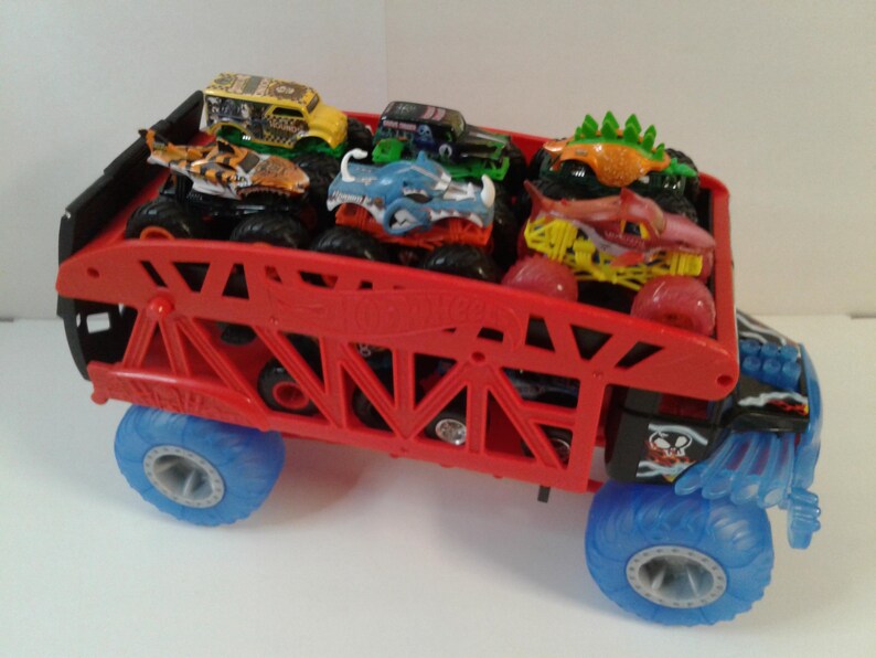 Może przedstawiać: Czerwona zabawka ciężar&oacute;wki potwora Hot Wheels przewożąca kilka mniejszych ciężar&oacute;wek potwor&oacute;w. Duża ciężar&oacute;wka ma niebieskie koła i czarno-czerwony korpus. Mniejsze ciężar&oacute;wki mają r&oacute;żne kolory i wzory.