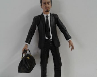 Peut inclure: Une figurine d'action noire et blanche d'un homme portant un costume noir et une cravate, tenant une mallette en cuir marron. La figurine a une expression sérieuse sur son visage.