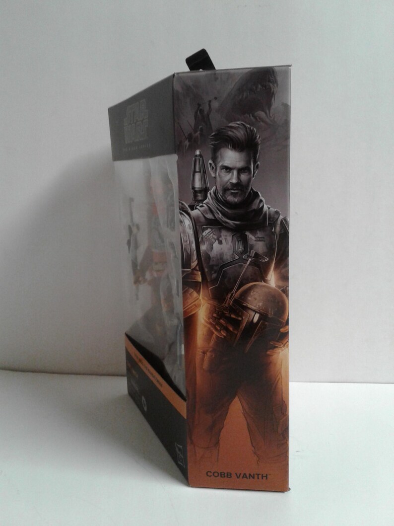 May include: A cardboard box with a Star Wars: The Mandalorian figure inside. The figure is of Cobb Vanth, a character from the show. The box is brown and has a picture of Cobb Vanth on the front. The text "COBB VANTH" is printed on the bottom of the box.