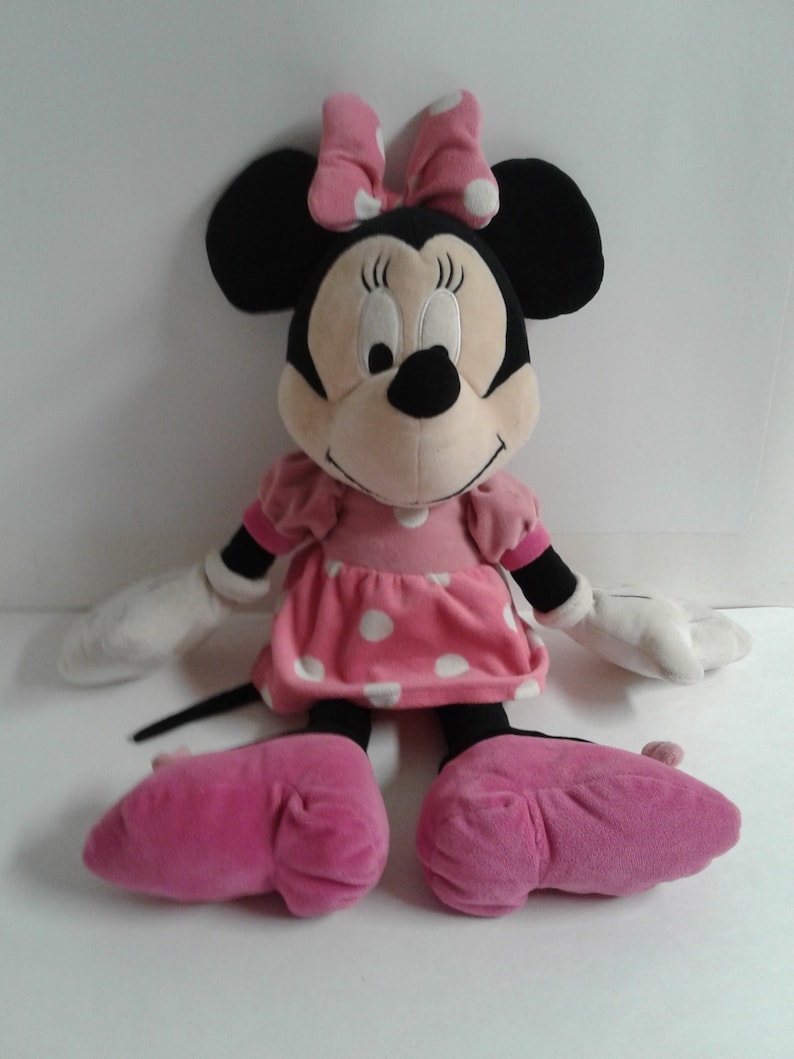 May include: A large plush Minnie Mouse toy wearing a pink polka dot dress and a pink bow. She has black ears and a black nose. Her eyes are closed and she is smiling.