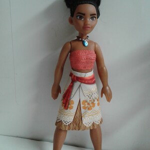 May include: A brown-skinned doll with black hair styled in a bun, wearing a red and white top, a white skirt with orange flowers, and a blue necklace.