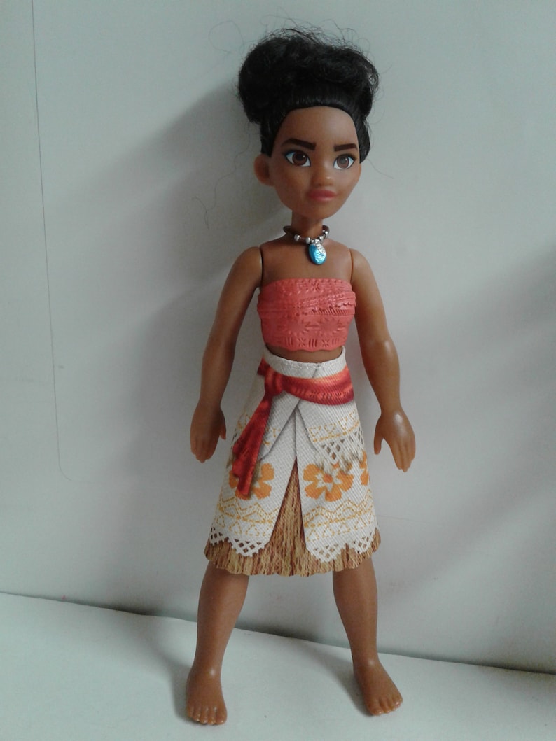 May include: A brown-skinned doll with black hair styled in a bun, wearing a red and white top, a white skirt with orange flowers, and a blue necklace.