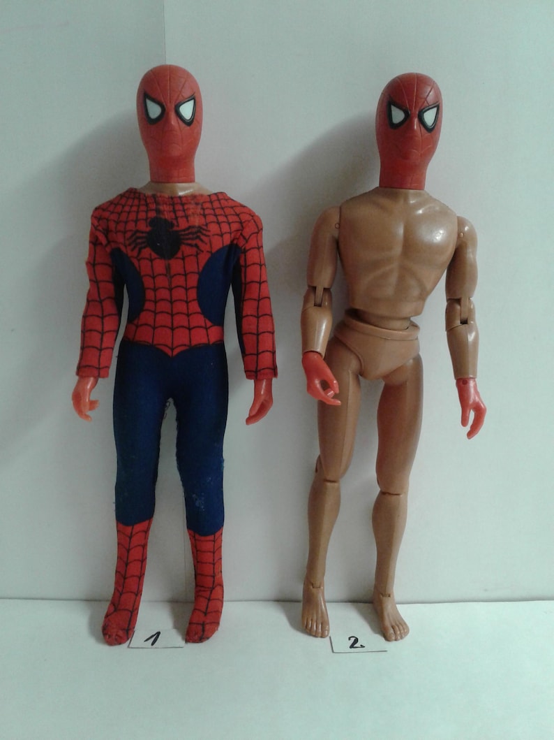 Op de afbeelding: Twee vintage action figuren van Spider-Man. De figuur links draagt een rood en blauw pak met een spinnenlogo op de borst. De figuur rechts is een naakt lichaam met een rood masker. Beide figuren hebben rode maskers met witte ogen.