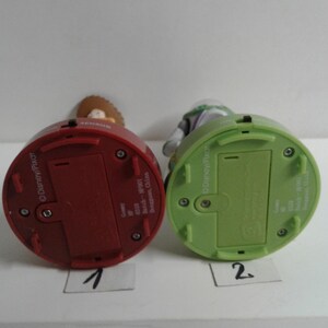 May include: Two Toy Story character figurines, one red and one green, are shown from the bottom. The red figurine is labeled "1" and the green figurine is labeled "2". The figurines have battery compartments and the Disney/Pixar logo.