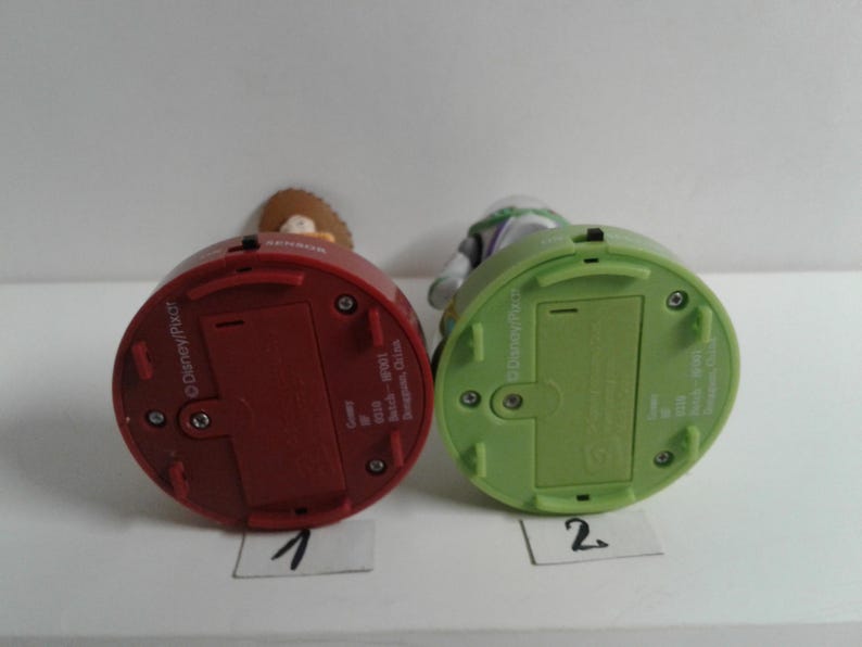 May include: Two Toy Story character figurines, one red and one green, are shown from the bottom. The red figurine is labeled "1" and the green figurine is labeled "2". The figurines have battery compartments and the Disney/Pixar logo.