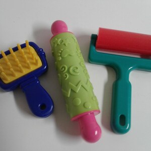 May include: Three plastic tools for play dough: a blue and yellow roller with spikes, a green rolling pin with a pink handle and a green and white patterned design, and a teal rolling pin with a red roller and a pink handle.